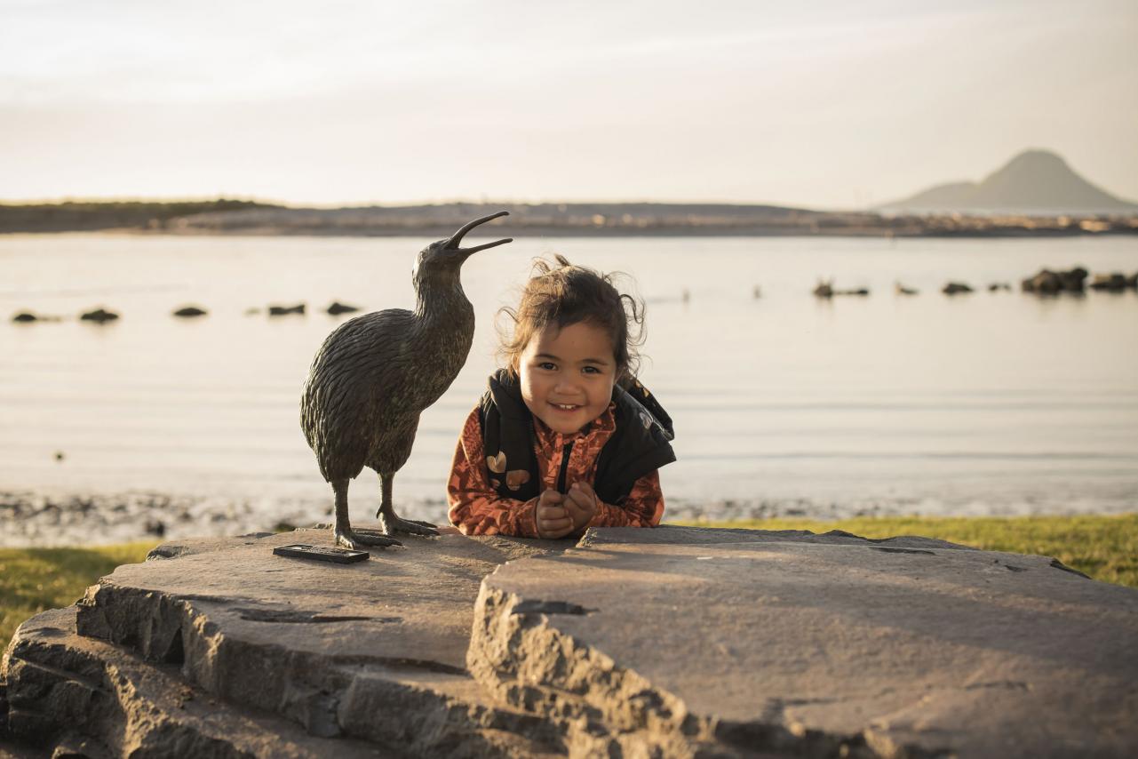 Child standing next to Kiwi statue with Moutohora in background